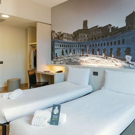 Modern hotel room with two twin beds, a large wall mural of ancient ruins, an open closet, and a small desk with a chair.