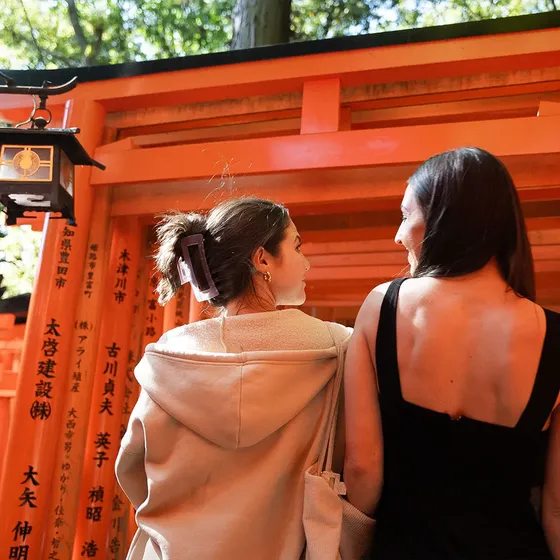 Two women stand facing each other in front of vibrant orange torii gates with Japanese inscriptions at a shrine. One wears a black dress, the other a hoodie.