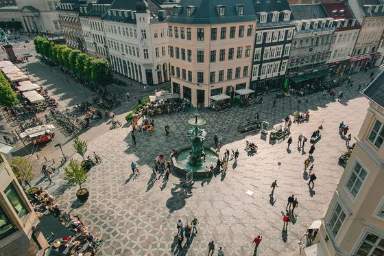 A city crossing featuring rows of brightly colored buildings with people walking around a fountain in the middle