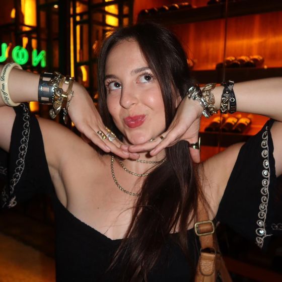 Woman posing playfully with hands under her chin, wearing multiple bracelets, a necklace, and an off-shoulder top in a warmly lit setting.