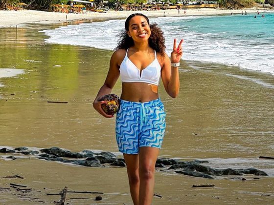 A woman in a white top and blue patterned shorts holds a coconut and flashes a peace sign on a tropical beach with lush greenery.
