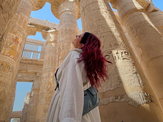 A person with red hair looks up at ancient, hieroglyph-covered stone columns in a sunlit historical site with a clear blue sky.