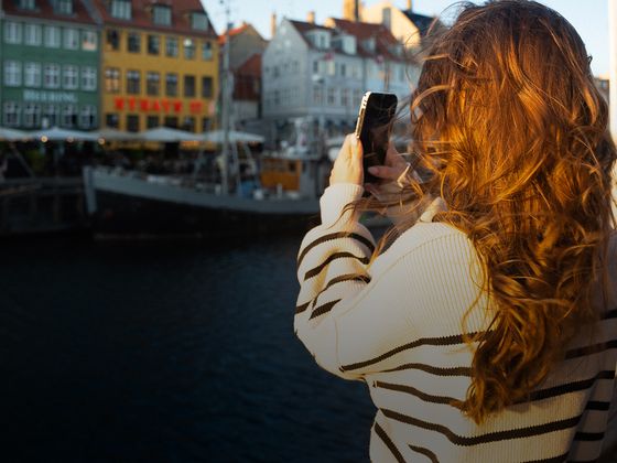 Woman with long hair takes a photo of colorful waterfront buildings at a harbor under a clear blue sky.
