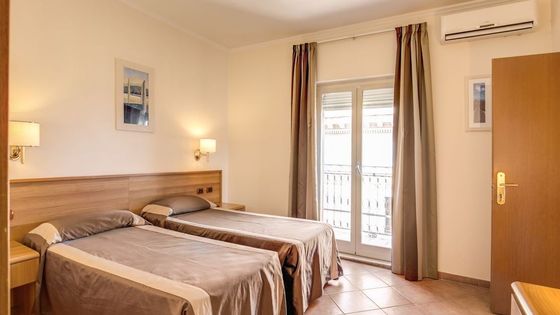 Hotel room with two single beds, wood headboard, wall lamps, framed art, curtains, air conditioner, and a door leading to a balcony.