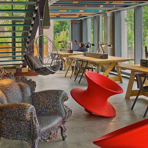 Modern open office space with colorful, eclectic furniture, hanging chair, and large windows. Features unique red chairs and patterned sofa.