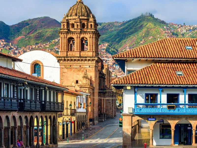 Historic city street with colonial buildings, red-tiled roofs, and a prominent stone tower against a backdrop of green mountains under a blue sky.