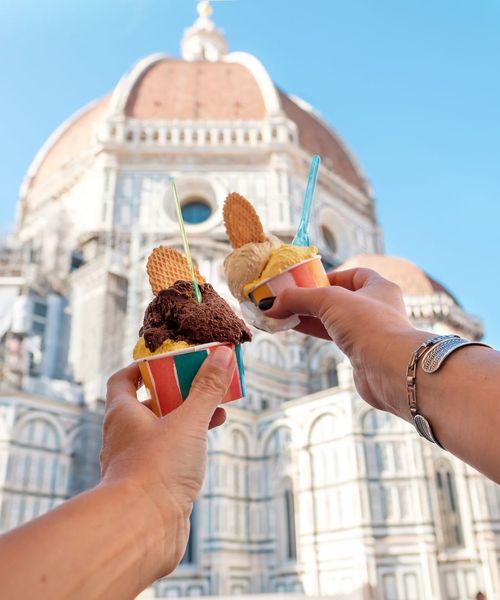 Two colorful cups of gelato with waffle pieces raised in front of a large domed building under a clear blue sky.
