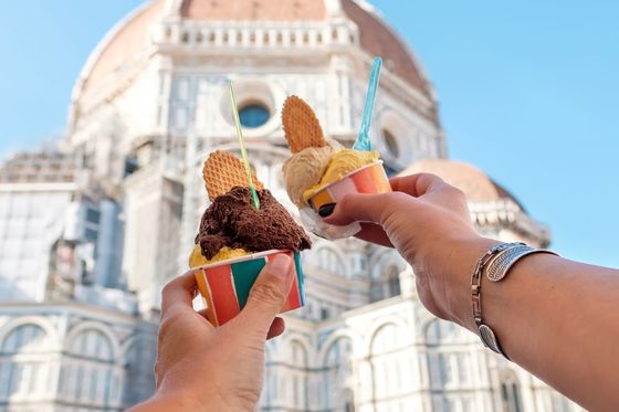 Two colorful cups of gelato with waffle pieces raised in front of a large domed building under a clear blue sky.