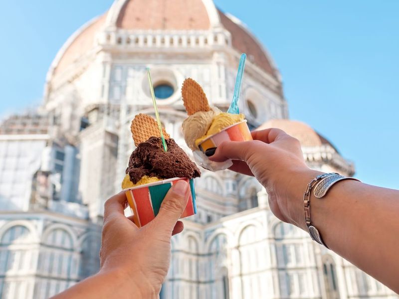 Two colorful cups of gelato with waffle pieces raised in front of a large domed building under a clear blue sky.