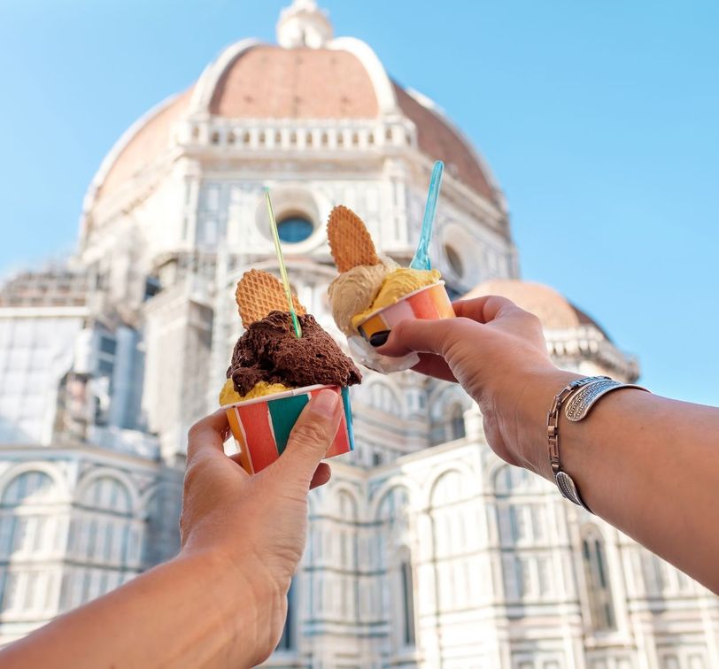 Two colorful cups of gelato with waffle pieces raised in front of a large domed building under a clear blue sky.