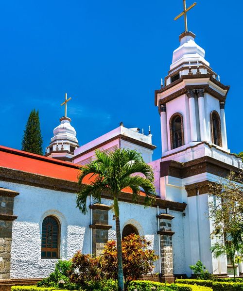 A white church with a red roof and two bell towers topped with crosses, surrounded by lush greenery under a clear blue sky.