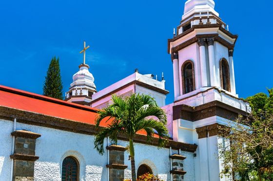 A white church with a red roof and two bell towers topped with crosses, surrounded by lush greenery under a clear blue sky.