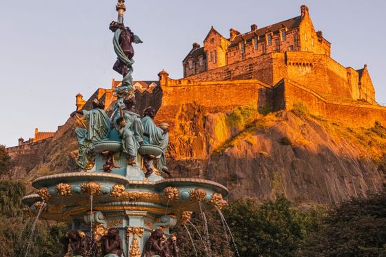 A historic stone castle on a rocky hill is illuminated by warm sunlight, with an ornate fountain in the foreground, surrounded by greenery.