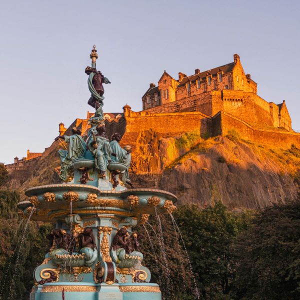 A historic stone castle on a rocky hill is illuminated by warm sunlight, with an ornate fountain in the foreground, surrounded by greenery.