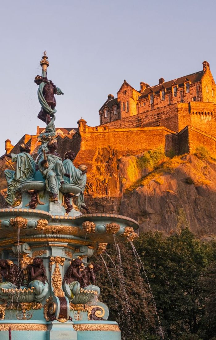 A historic stone castle on a rocky hill is illuminated by warm sunlight, with an ornate fountain in the foreground, surrounded by greenery.