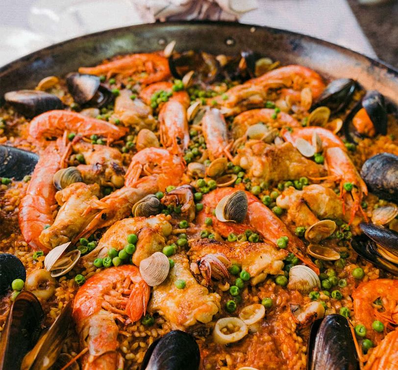 A pan of seafood paella with prawns, mussels, clams, green peas, and rice, garnished with herbs, displayed on a table.