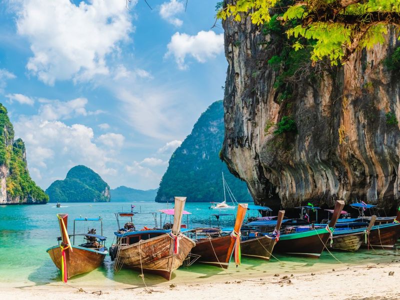 Longtail boats docked on a sandy beach beneath a rocky cliff, with vibrant green foliage and blue ocean stretching to distant islands under a clear sky.