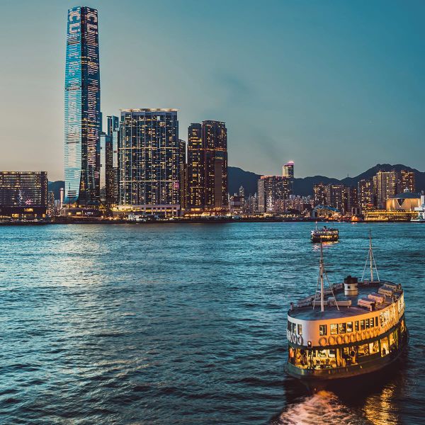 Ferry sailing in a harbor at twilight, with a city skyline and a tall skyscraper in the background.