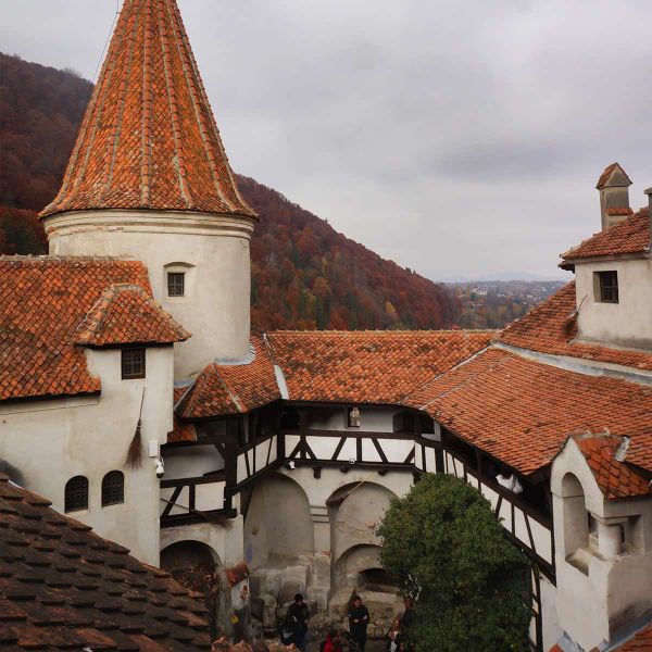 Castle with orange-tiled roofs set against forested hills under a cloudy sky. Medieval architecture with visible archways and turrets.