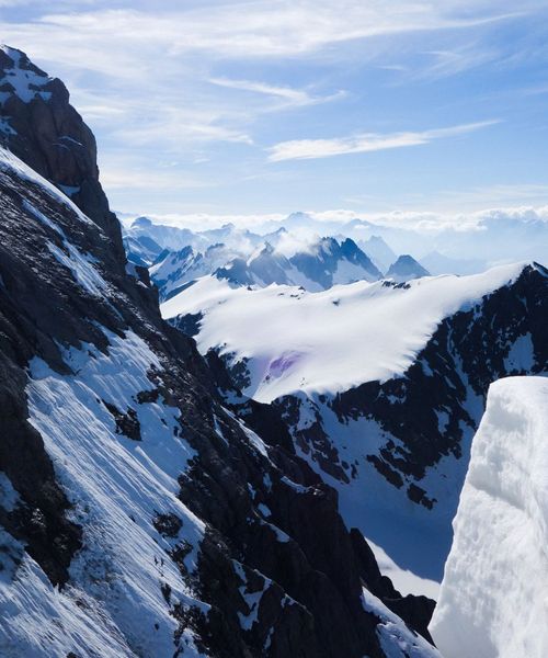 Snow-covered mountain peaks under a clear blue sky, with rugged black rock faces and distant foggy mountains in the background.
