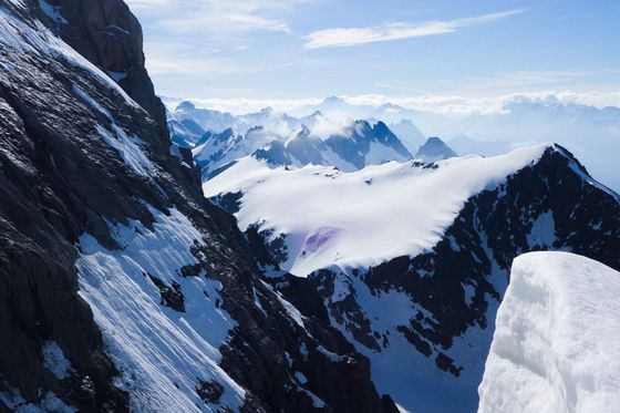 Snow-covered mountain peaks under a clear blue sky, with rugged black rock faces and distant foggy mountains in the background.