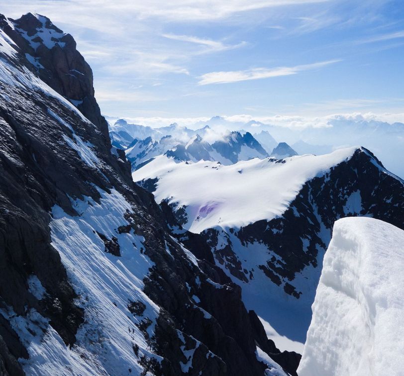 Snow-covered mountain peaks under a clear blue sky, with rugged black rock faces and distant foggy mountains in the background.