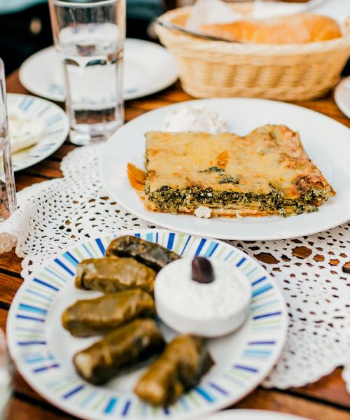 A table with a plate of spinach pie and a dish of dolmas with a small bowl of yogurt, surrounded by glasses of water and bread in a basket.
