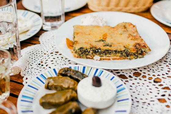 A table with a plate of spinach pie and a dish of dolmas with a small bowl of yogurt, surrounded by glasses of water and bread in a basket.