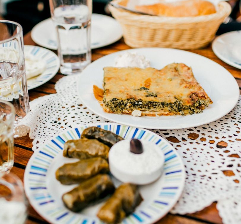 A table with a plate of spinach pie and a dish of dolmas with a small bowl of yogurt, surrounded by glasses of water and bread in a basket.