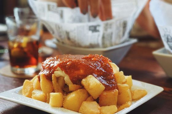 A plate of patatas bravas topped with red sauce and aioli on a wooden table, with a hand reaching into a basket of bread in the background.