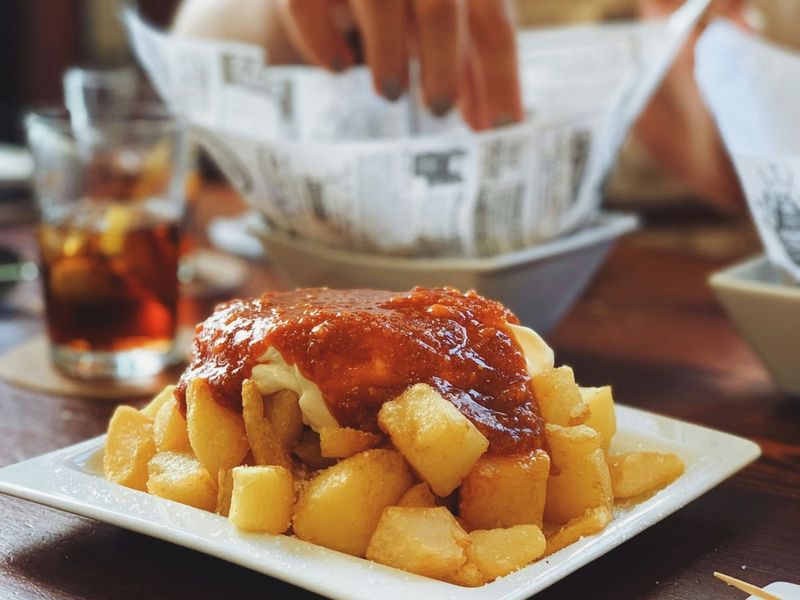A plate of patatas bravas topped with red sauce and aioli on a wooden table, with a hand reaching into a basket of bread in the background.