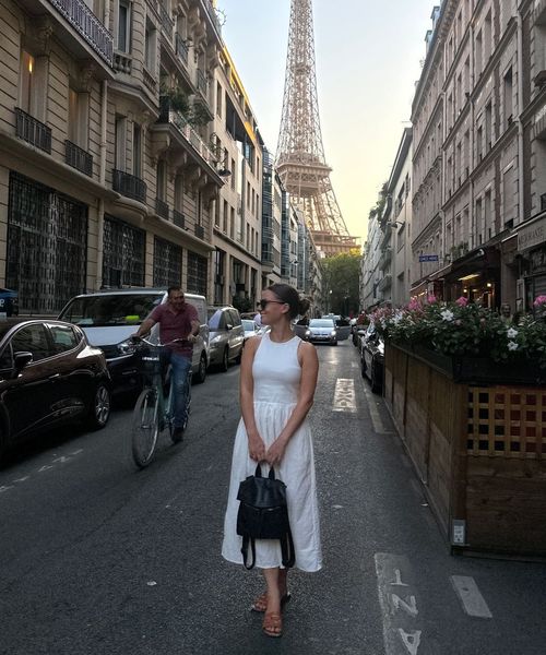 A woman in a white dress stands on a Parisian street with the Eiffel Tower in the background, surrounded by cars and a cyclist.