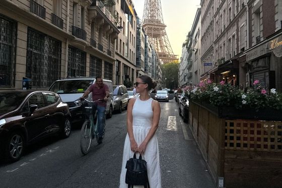A woman in a white dress stands on a Parisian street with the Eiffel Tower in the background, surrounded by cars and a cyclist.