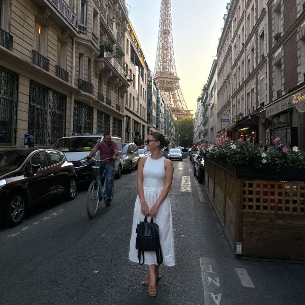 A woman in a white dress stands on a Parisian street with the Eiffel Tower in the background, surrounded by cars and a cyclist.