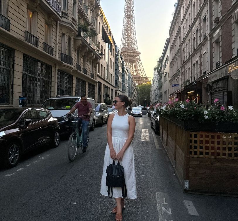 A woman in a white dress stands on a Parisian street with the Eiffel Tower in the background, surrounded by cars and a cyclist.