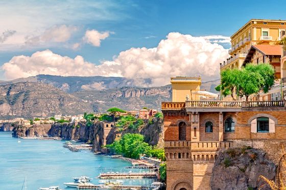 Cliffside buildings overlook a marina with boats docked along the turquoise coastline, set against a backdrop of mountains and a partly cloudy sky.