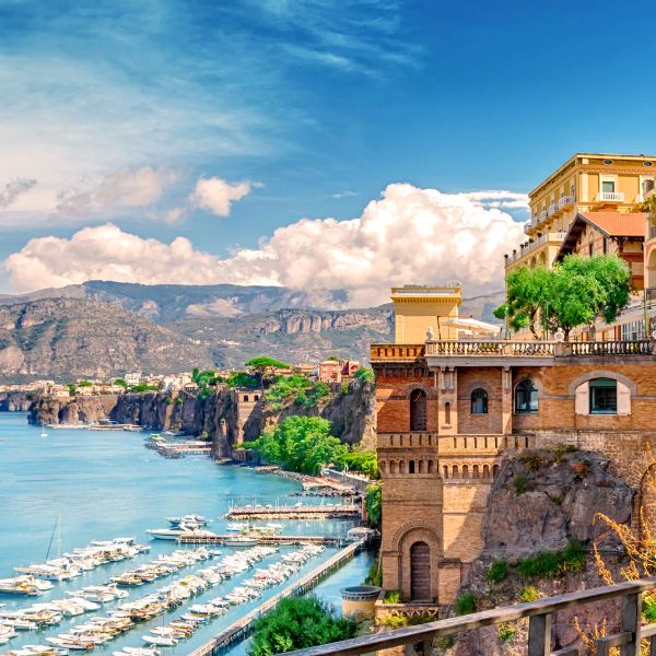 Cliffside buildings overlook a marina with boats docked along the turquoise coastline, set against a backdrop of mountains and a partly cloudy sky.