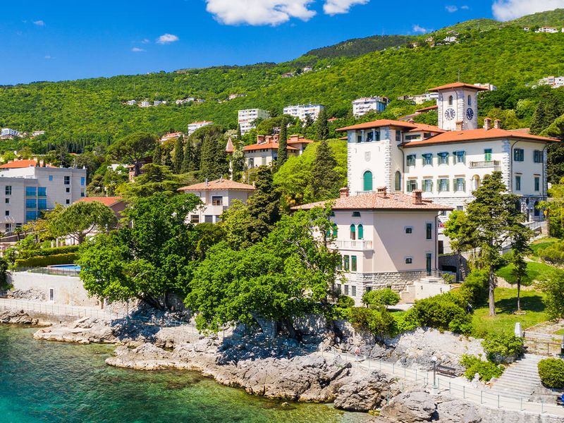 Coastal landscape with historic buildings surrounded by greenery, clear turquoise water, and a hillside dotted with trees and houses under a blue sky.
