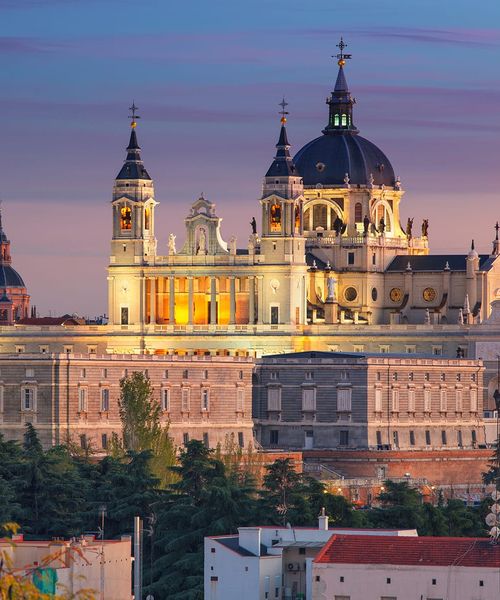 Illuminated Almudena Cathedral in Madrid at dusk, featuring ornate architecture and a large central dome against a colorful evening sky.