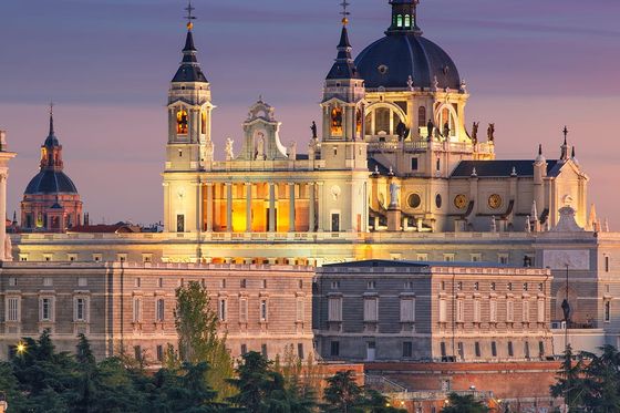 Illuminated Almudena Cathedral in Madrid at dusk, featuring ornate architecture and a large central dome against a colorful evening sky.