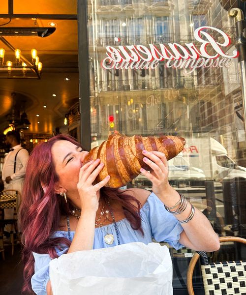 A woman playfully biting a giant croissant outside a French cafe, with a stylish interior visible behind her and reflections in the cafe window.