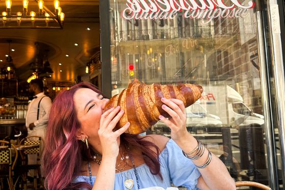 A woman playfully biting a giant croissant outside a French cafe, with a stylish interior visible behind her and reflections in the cafe window.
