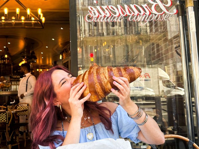 A woman playfully biting a giant croissant outside a French cafe, with a stylish interior visible behind her and reflections in the cafe window.