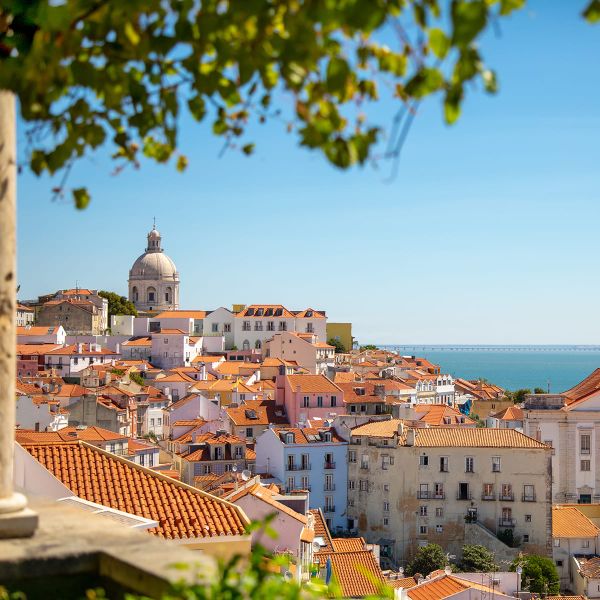 Scenic view of Lisbon with red-roofed buildings, a dome, and the sea in the background, framed by green leaves.