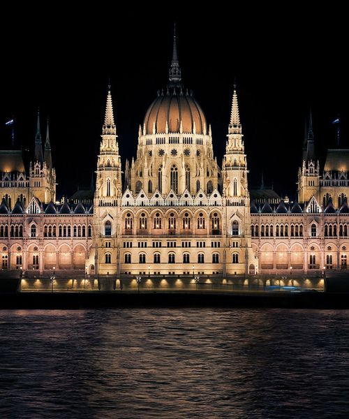 Illuminated gothic-style building with dome and spires, reflecting on a river at night.
