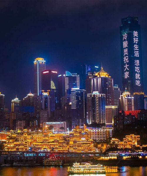Illuminated cityscape at night with skyscrapers and brightly lit buildings reflecting on a river, featuring vertical Chinese text on a tall tower.