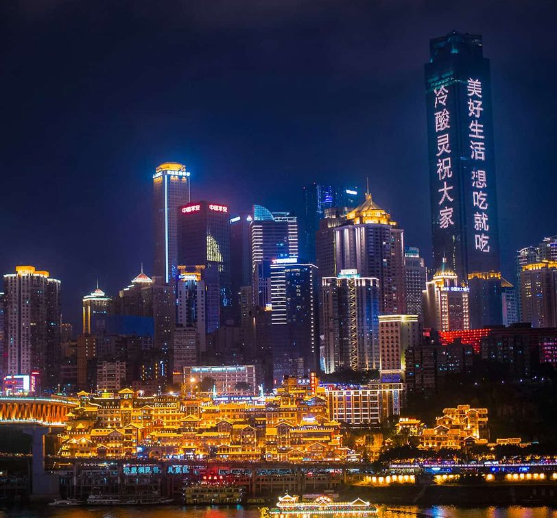 Illuminated cityscape at night with skyscrapers and brightly lit buildings reflecting on a river, featuring vertical Chinese text on a tall tower.