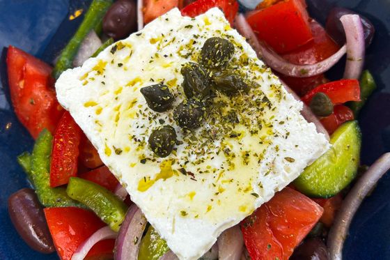 A Greek salad with feta cheese topped with capers, surrounded by tomatoes, cucumbers, red onions, bell peppers, and olives in a blue bowl.
