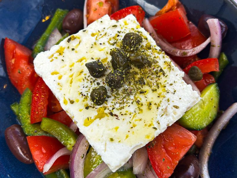 A Greek salad with feta cheese topped with capers, surrounded by tomatoes, cucumbers, red onions, bell peppers, and olives in a blue bowl.