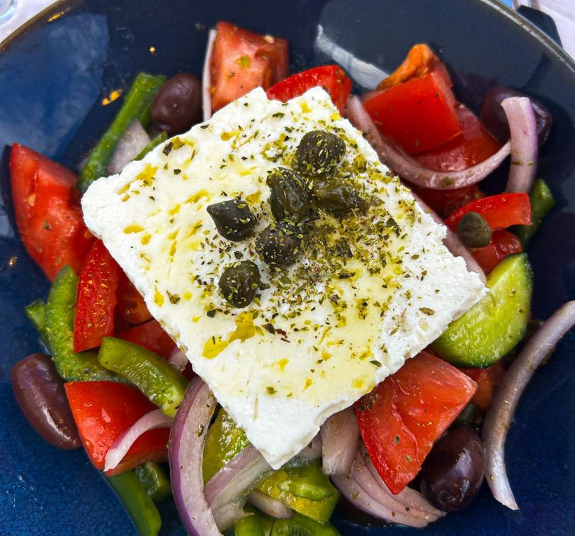 A Greek salad with feta cheese topped with capers, surrounded by tomatoes, cucumbers, red onions, bell peppers, and olives in a blue bowl.